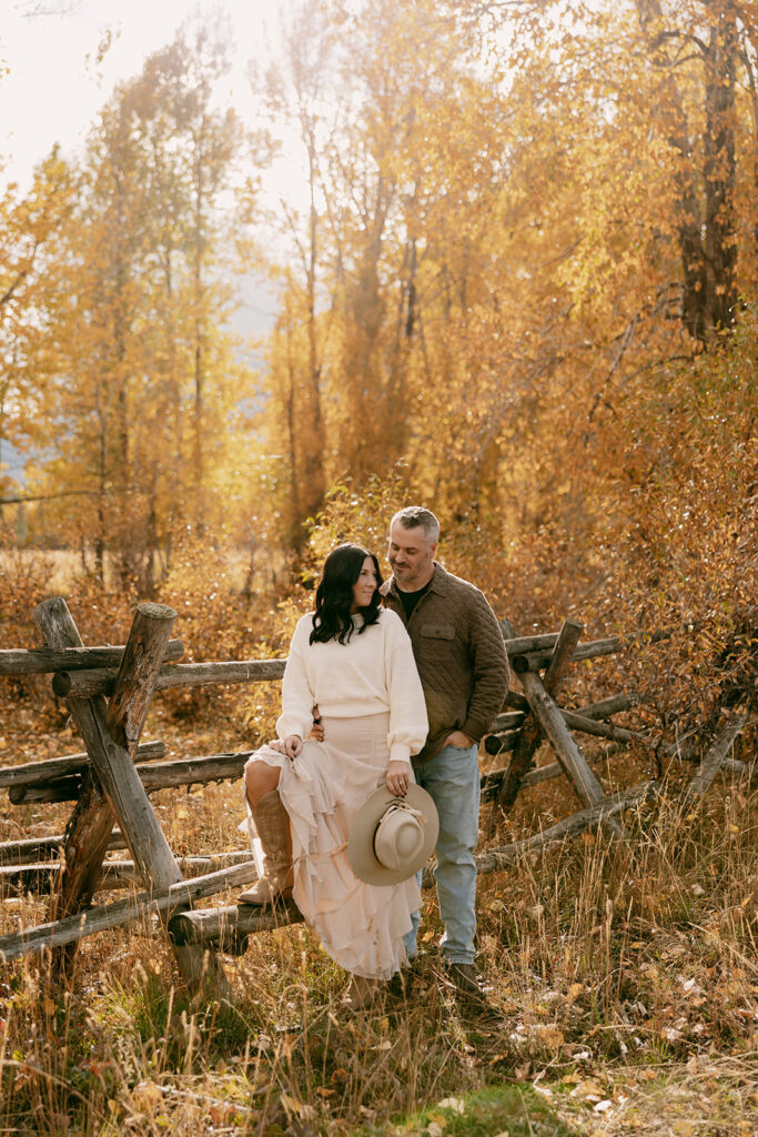 Engaged couple posing near rustic wooden fence under golden aspens during their Grand Tetons engagement photography session.