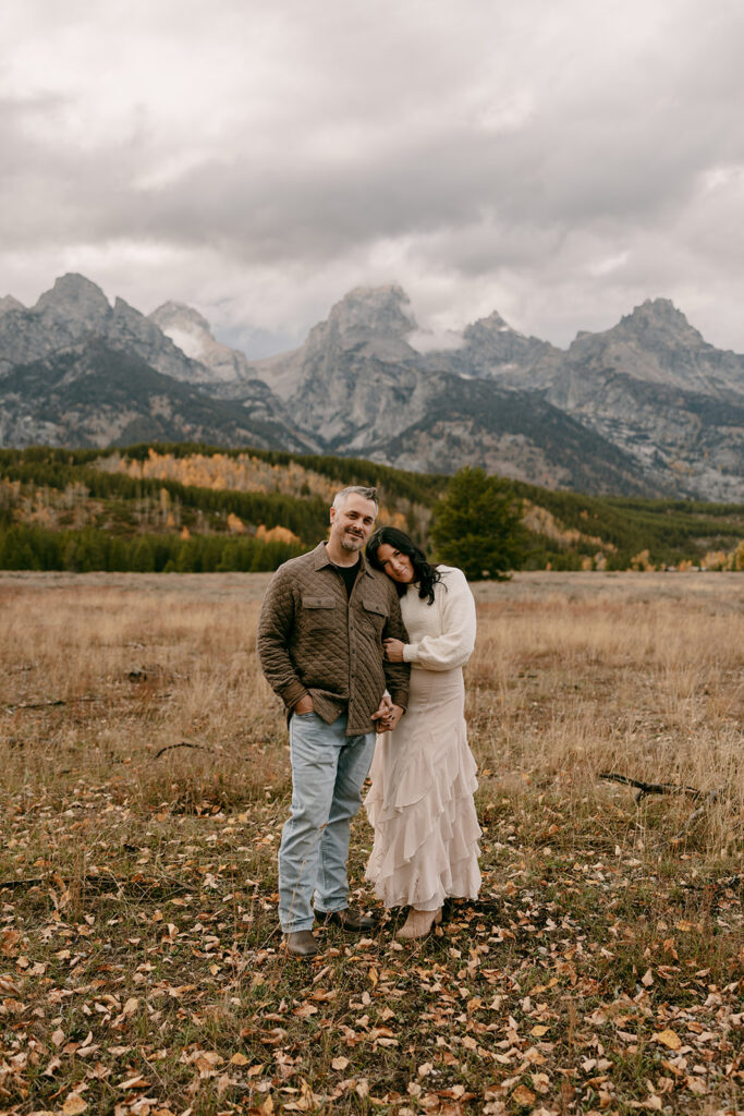 Engaged couple standing in a scenic meadow with dramatic Teton mountain peaks in the background during fall photography session.