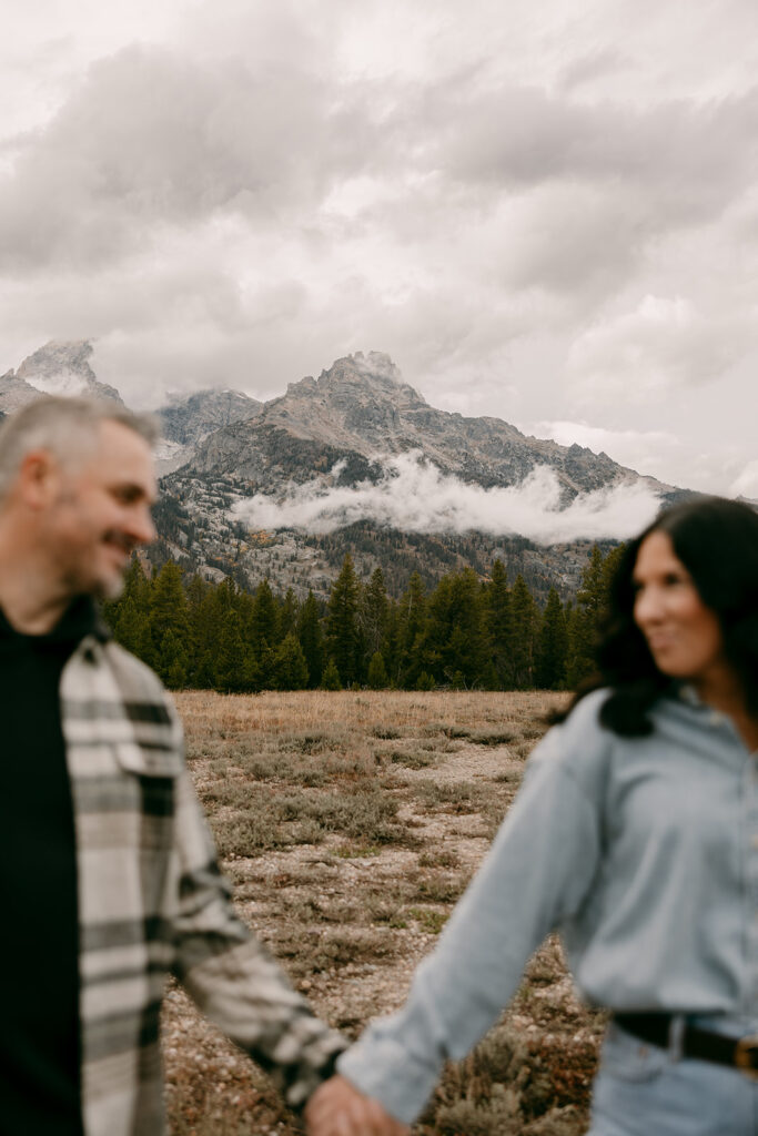 Cinematic Grand Tetons engagement photography shot with mountains in focus and couple holding hands in soft foreground.