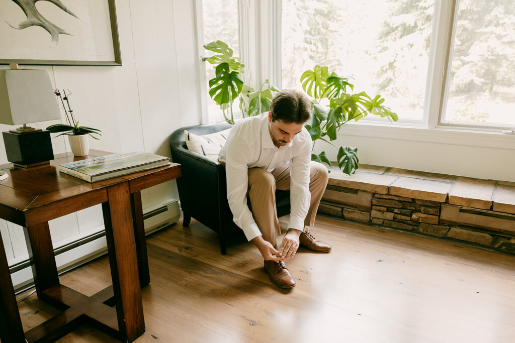 Groom ties brown dress shoes in a cozy lodge living room with natural light and forest views.