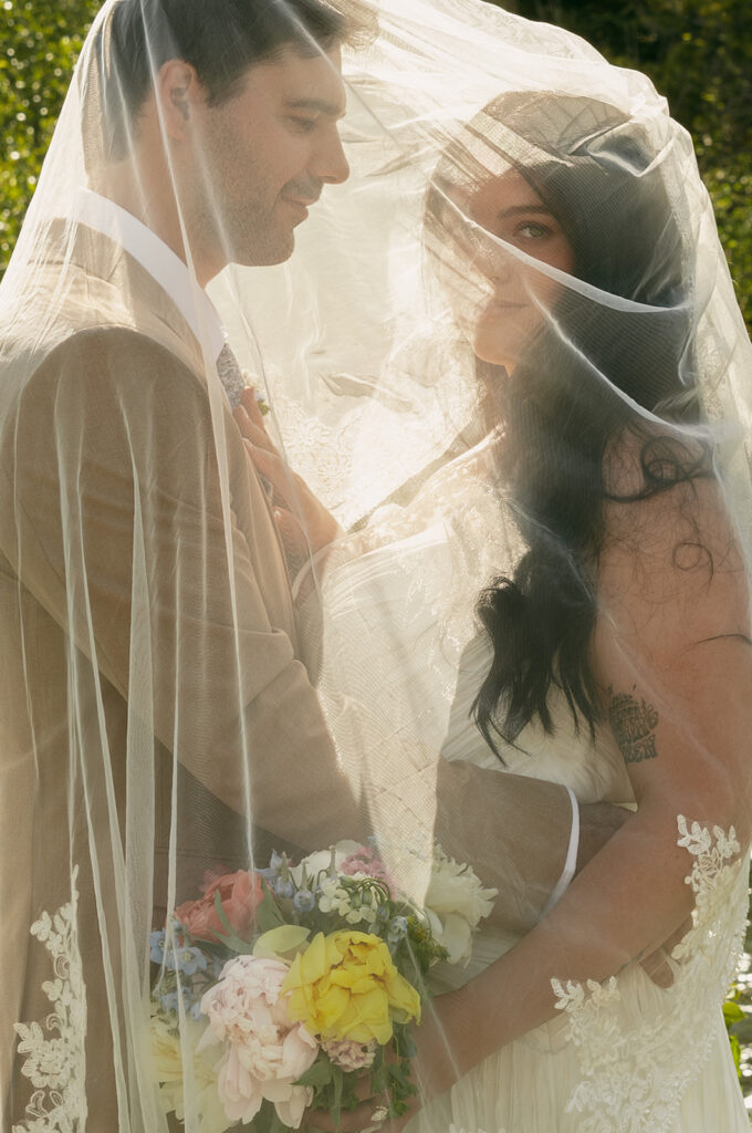 Bride and groom smile beneath a delicate lace-edged veil with sunlight filtering through the fabric.