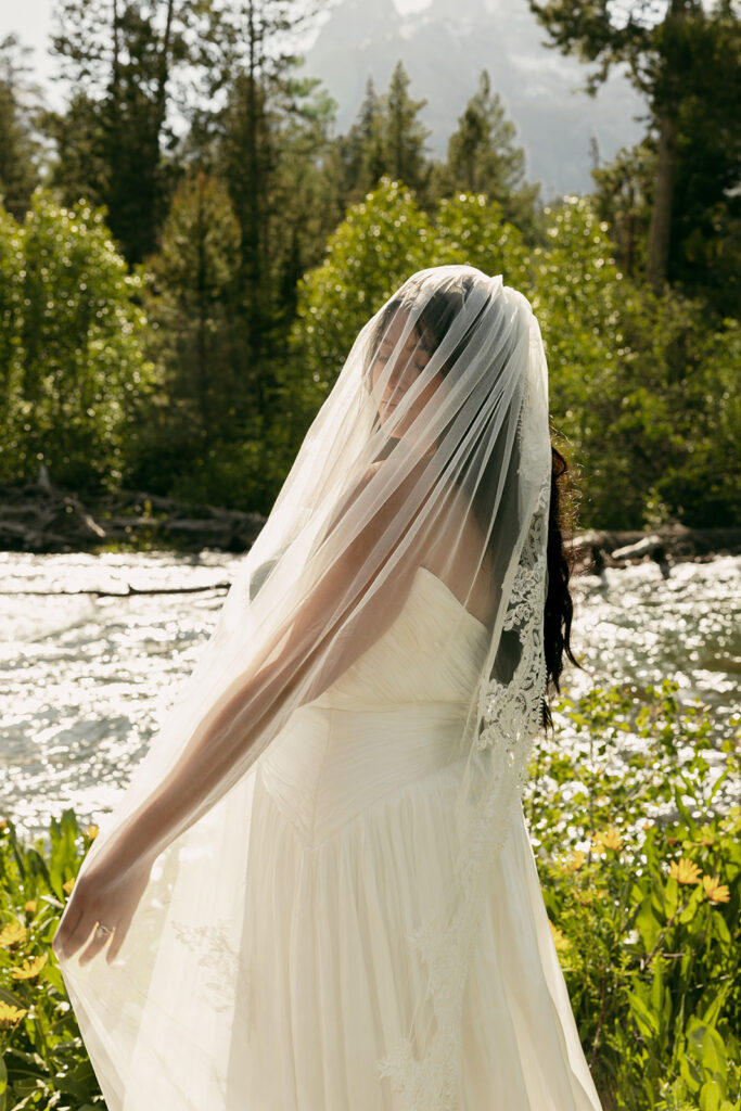 Bride stands by a sparkling riverside in the Grand Tetons, her veil draped over her body.