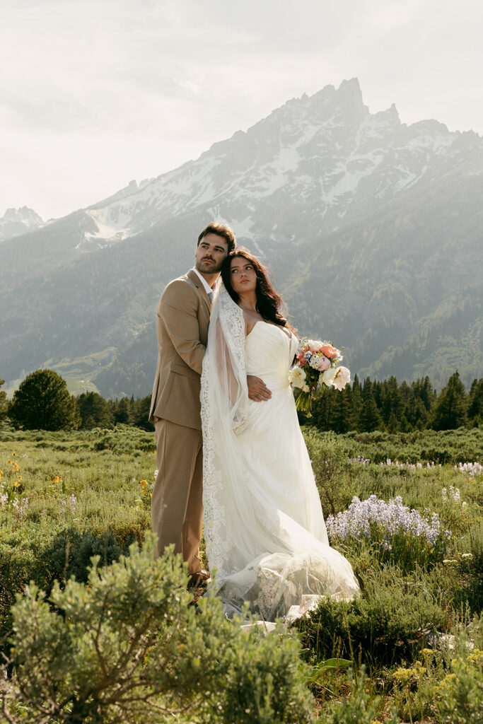 Bride and groom embracing in a mountain meadow with the Grand Tetons rising behind them.