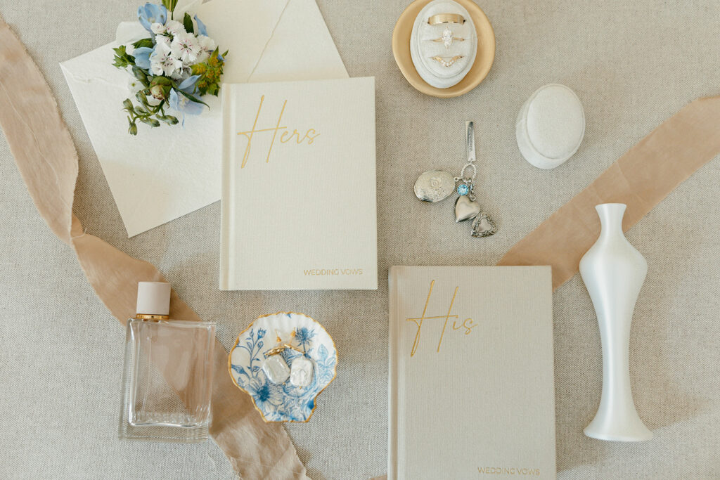 Flat lay of wedding details featuring vow books, rings, perfume, heirlooms, and soft florals.