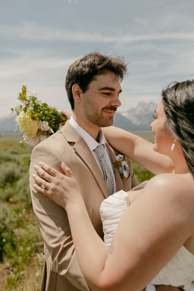 Groom smiles as bride embraces him, bouquet resting on his shoulder, with Grand Teton peaks in soft focus.
