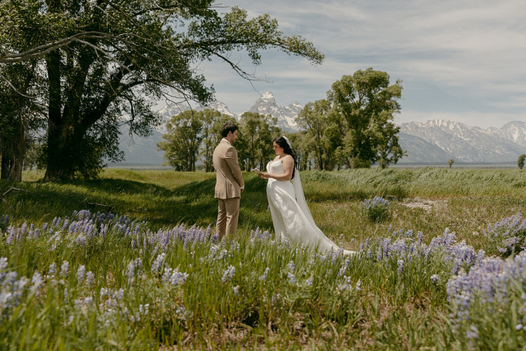Couple reads vows beneath trees and wildflowers, with snow-capped mountains creating a cinematic backdrop for this intimate wedding in the Grand Tetons.