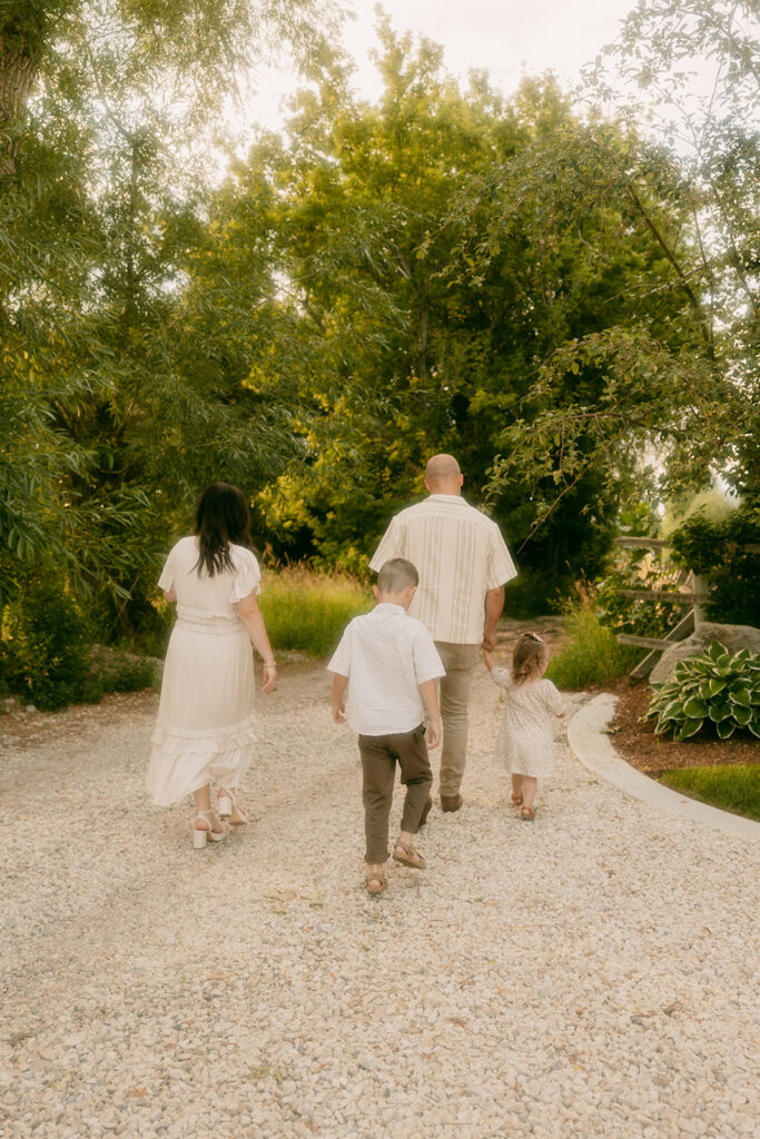 A family of four walking away from Idaho Falls family photographer surrounded by green trees and golden sunlight.