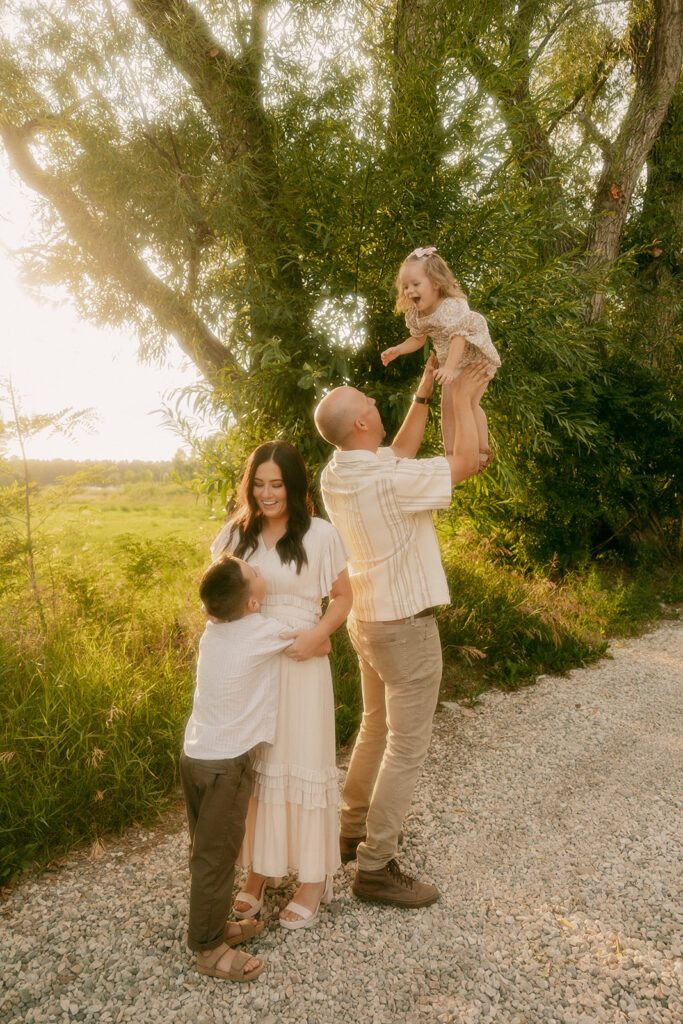 Dad lifts daughter playfully into the air as mom smiles, hugging their son beside her in a sunlit grove.
