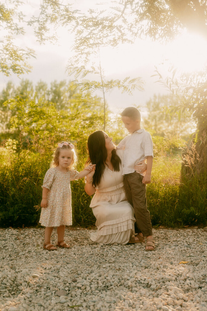 Mother kneels on a gravel path smiling at her two children, holding their hands as warm light filters through the trees.