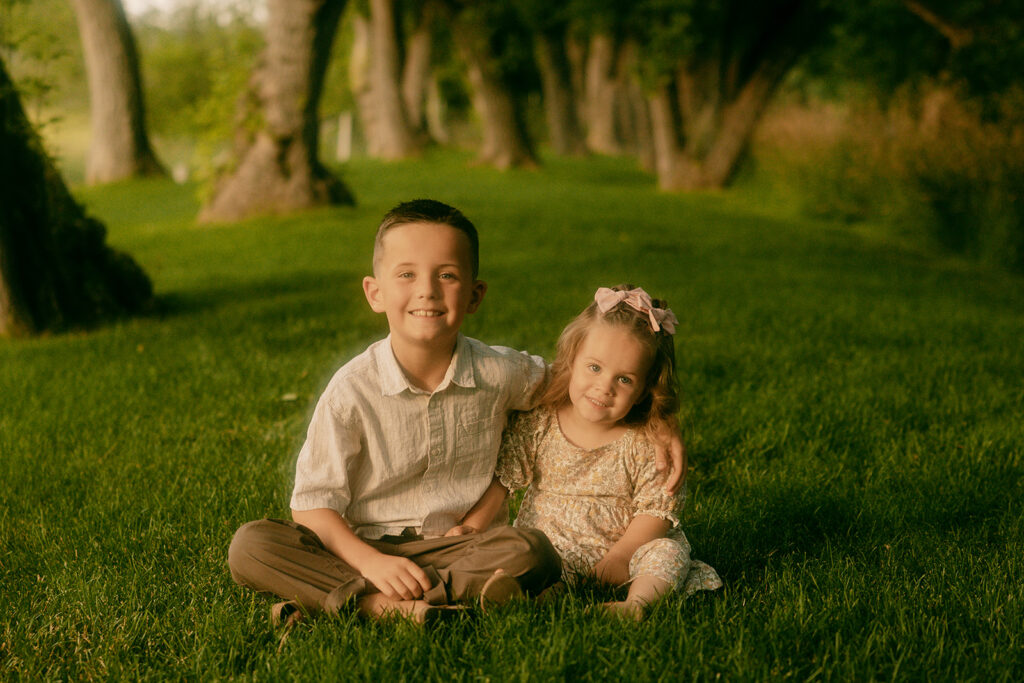 Brother and sister sitting close together in the grass, smiling at the camera under a canopy of trees.
