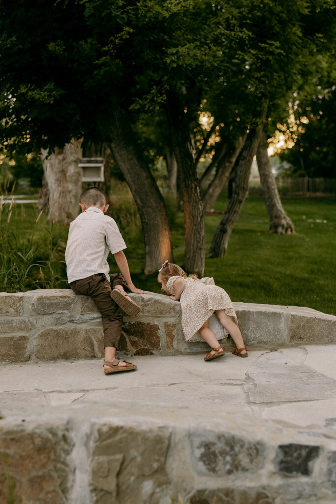 Brother and sister climbing on a stone wall under tall trees during golden hour playtime.