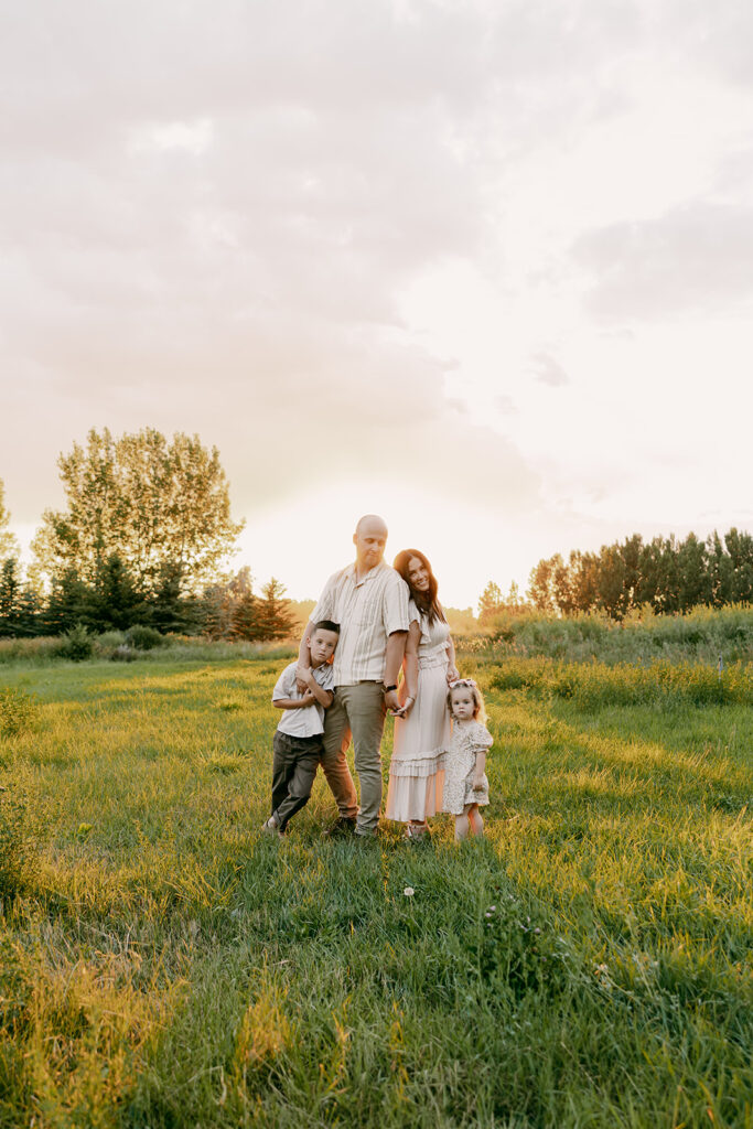 Family of four standing in tall grass, holding hands during a golden hour Idaho Falls family photographer session.