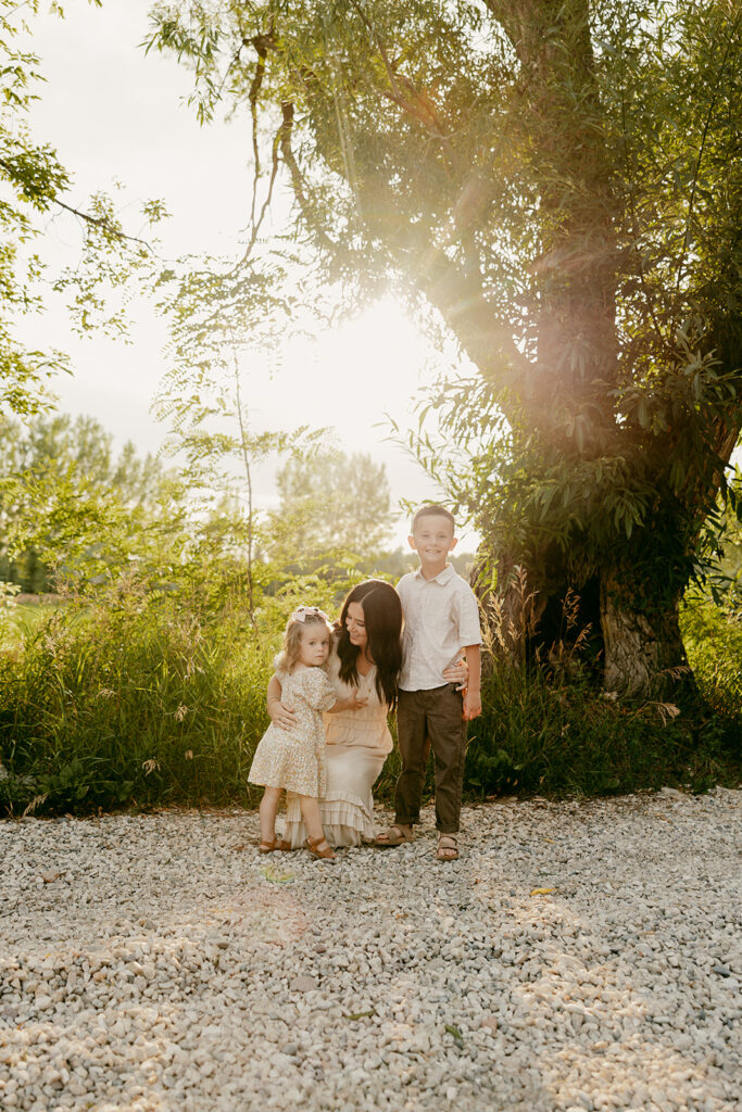 Mom with daughter and son near a tree-lined gravel path lit by golden sunlight.