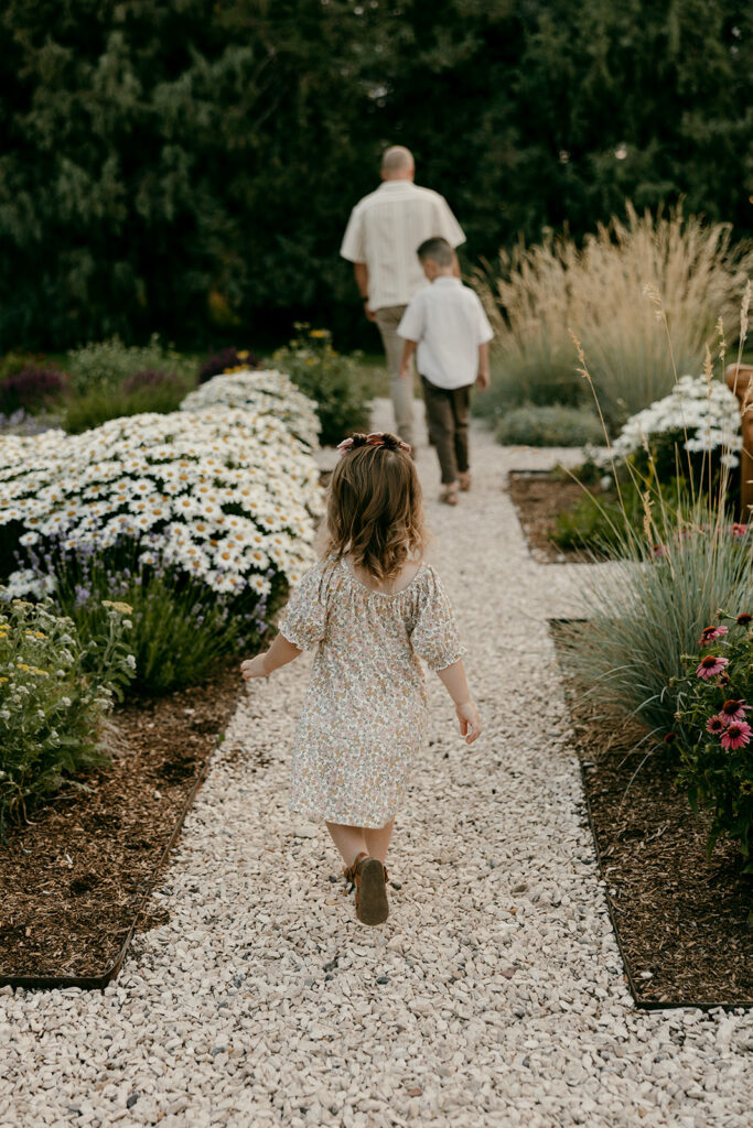 Toddler girl walking down a gravel garden path behind her father and brother.