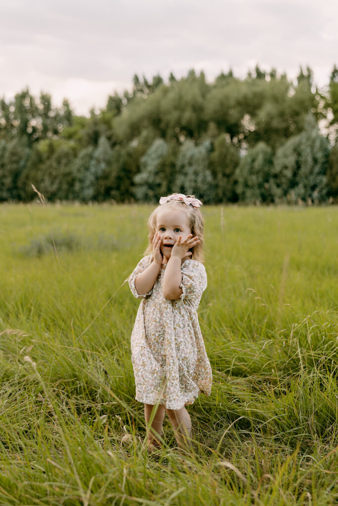 Toddler girl holding her cheeks with both hands, playing in a grassy field.