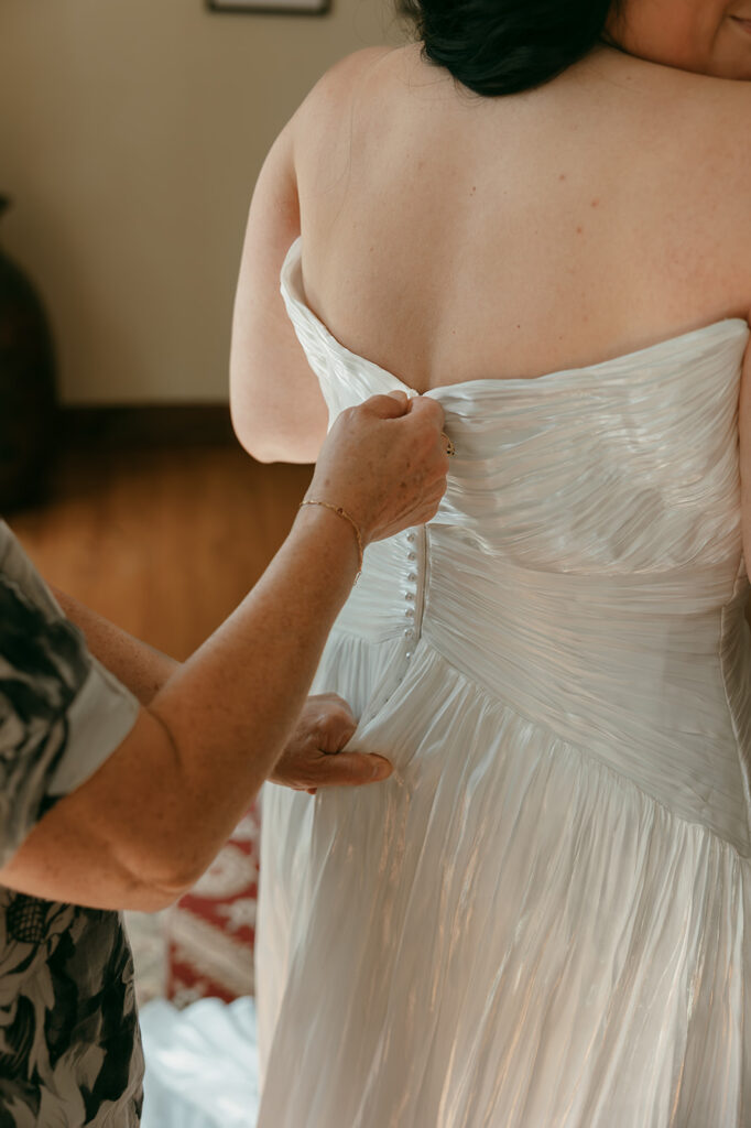 Mother zips the bride’s textured white gown during indoor getting-ready moments.