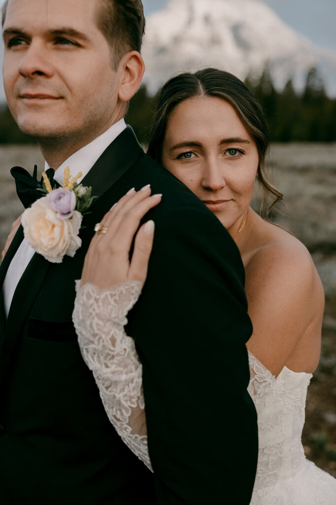Bride looks into camera while hugging groom from behind with Teton mountains in background.